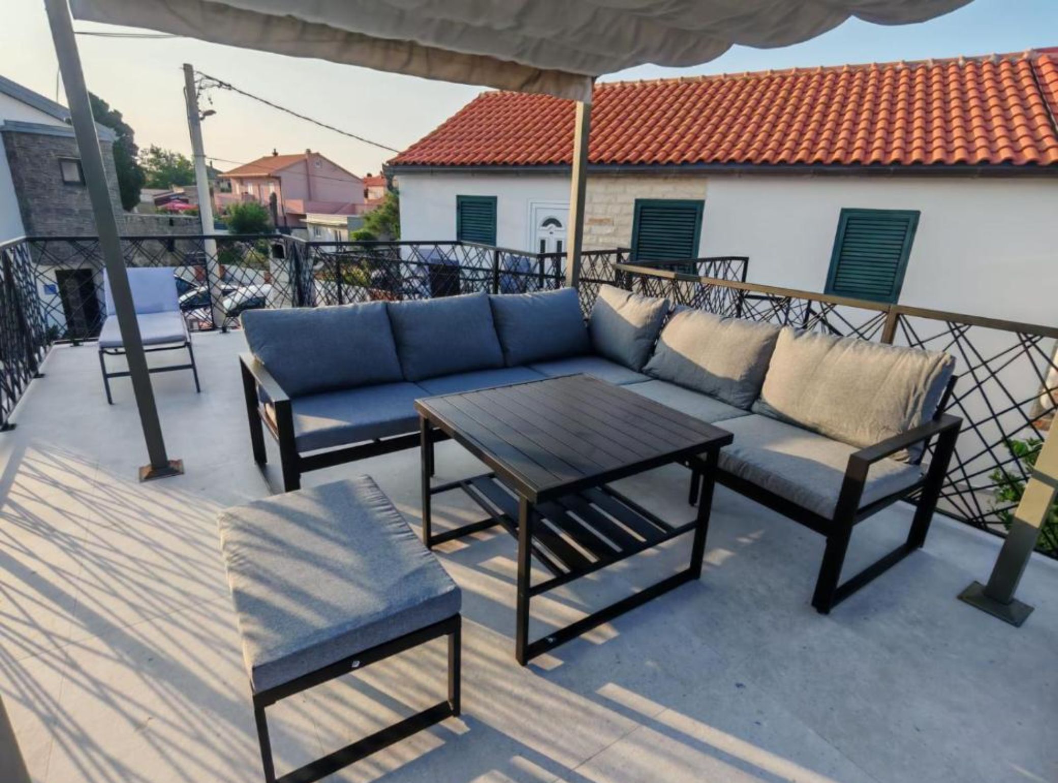 An outdoor seating area with a sofa and a table, a metal fence, and a building with a red-tiled roof in the background.