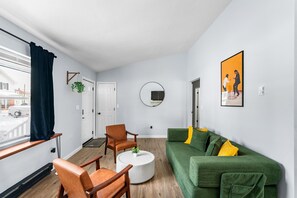 Una sala de estar moderna con un sofá verde, sillas de madera, una mesa de centro blanca y un gran ventanal con cortina.