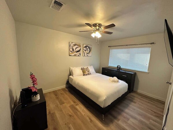 A bedroom with a bed, a ceiling fan, a window with blinds, and a small table with a plant.