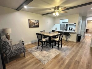 Un comedor con una mesa y sillas de madera, un ventilador de techo y una cocina al fondo.