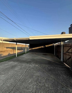 Un área de estacionamiento techada con piso de concreto y cerca de madera, bajo un cielo despejado.
