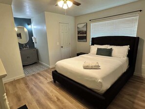 A bedroom with a bed, a ceiling fan, a window with blinds, a door, and a small dresser.