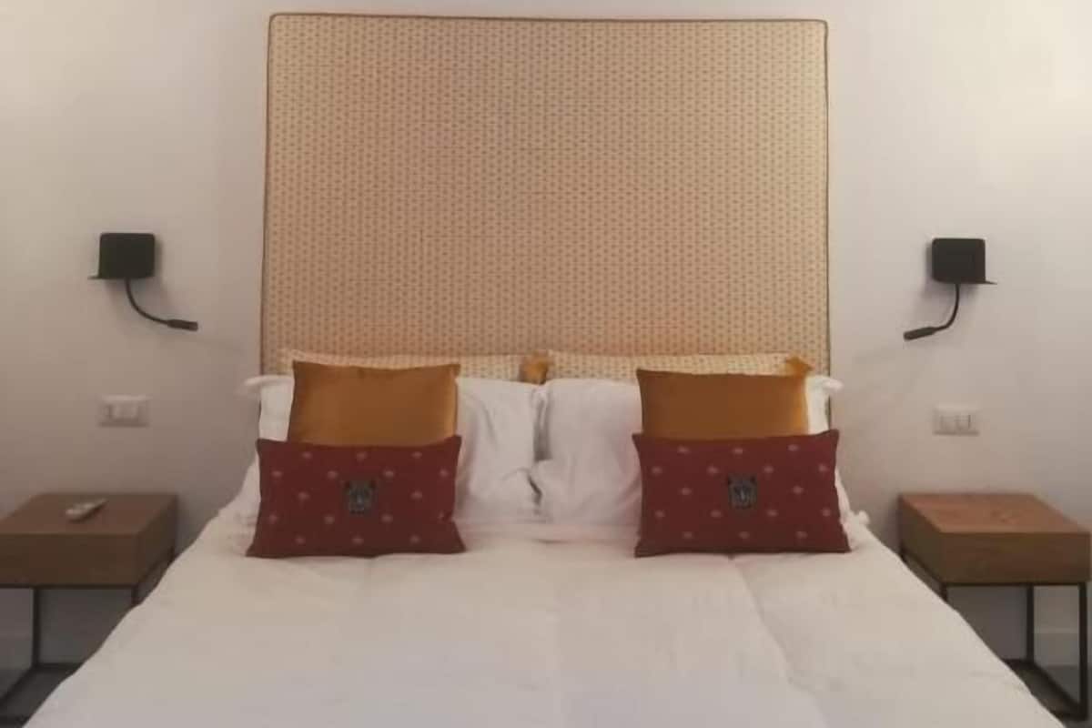 A bedroom with a large bed, two bedside tables, and a plain wall with a textured headboard.