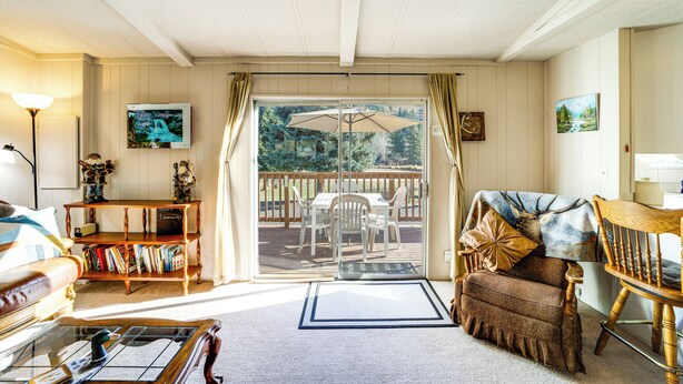 A cozy living room with a sofa, a coffee table, a bookshelf, and a view of a deck with outdoor furniture. - Cozy Waterfront Home w/ Mtn View Near Leavenworth (Peshastin)