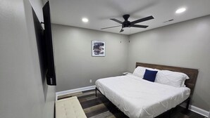 Un dormitorio con cama, ventilador de techo, mesita de noche pequeña y televisor de pared.