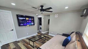 Un dormitorio con cama, televisor montado en la pared, ventilador de techo y lavadora al fondo.