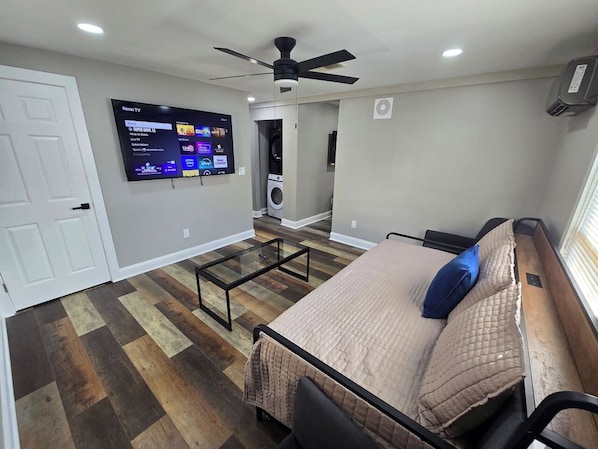 A bedroom with a bed, a TV mounted on the wall, a ceiling fan, and a washing machine in the background.