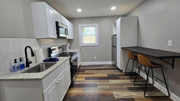 Una cocina compacta con armarios blancos, un lavamanos de acero inoxidable, un microondas y un refrigerador. El piso es de madera y hay una pequeña zona de comedor con una mesa y dos sillas.