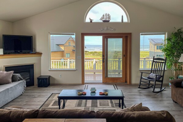 Una sala de estar con chimenea, televisor, una mesa de centro con adornos y vista al océano a través de amplias ventanas.
