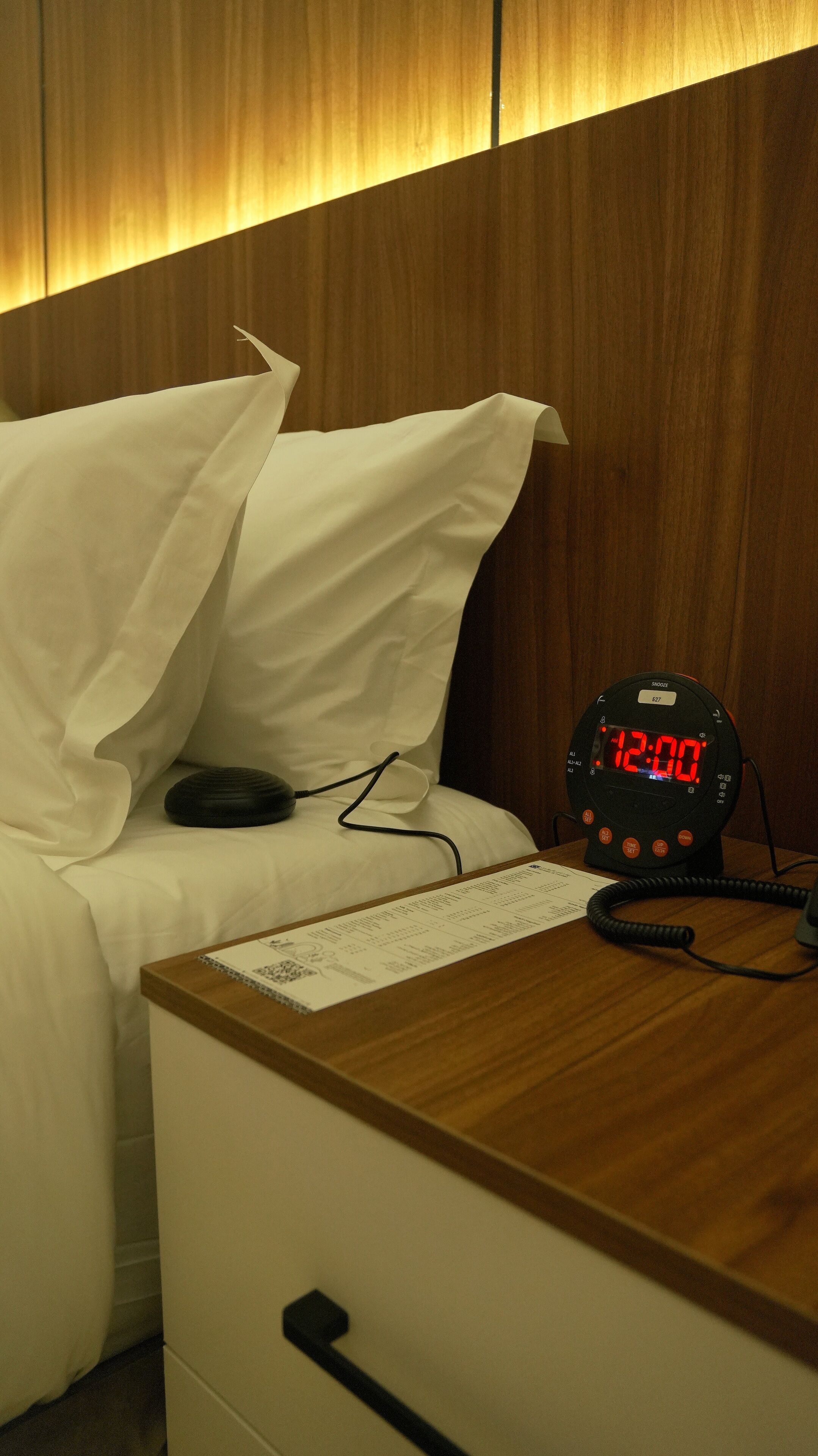 A hotel room with a wooden headboard, a bed with white bedding, a bedside table with a clock displaying 12:00, and a notepad on the table.