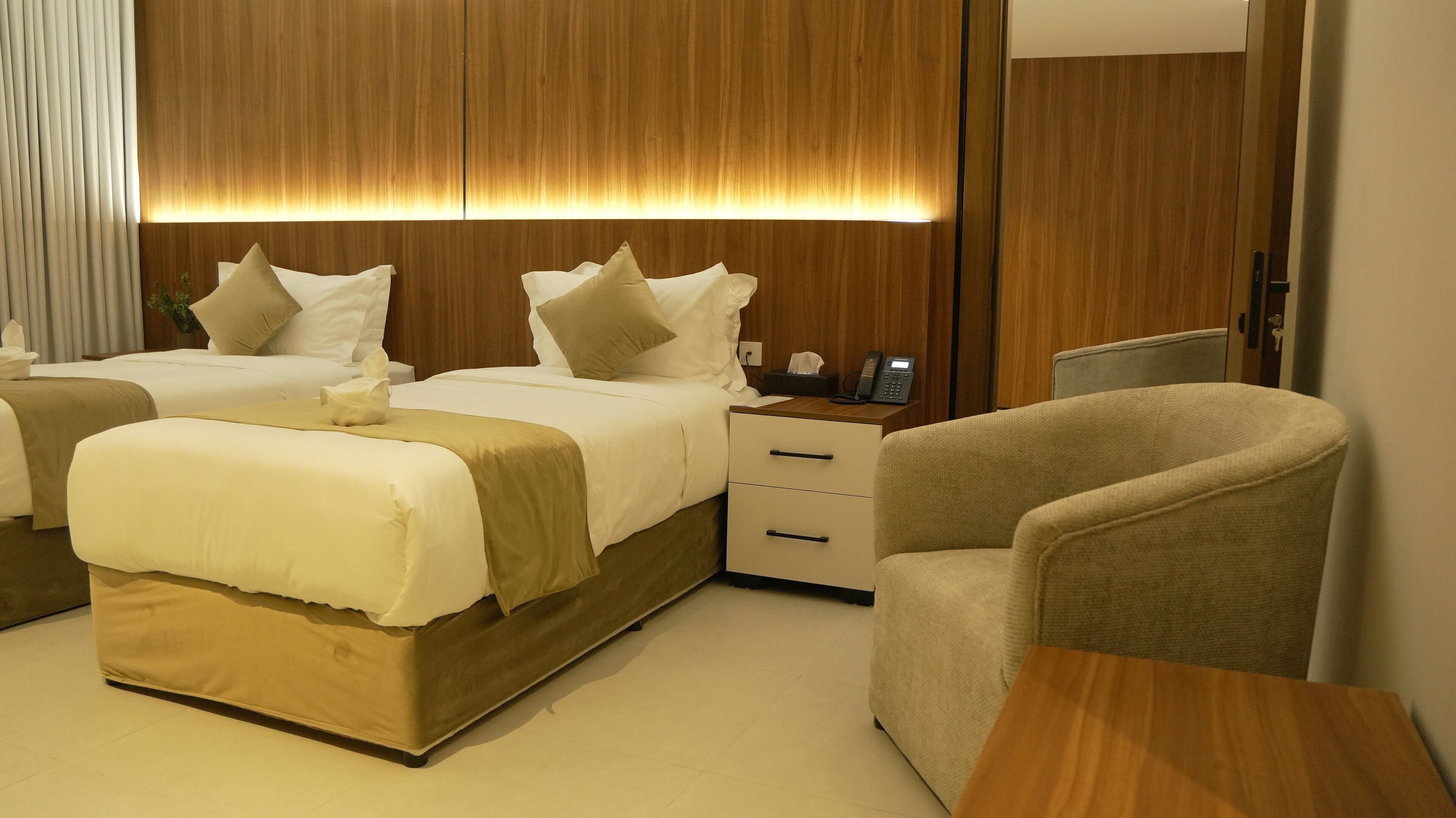 Habitación de hotel con dos camas, una mesita de noche con teléfono, una silla y un cabecero de madera con iluminación ambiental.