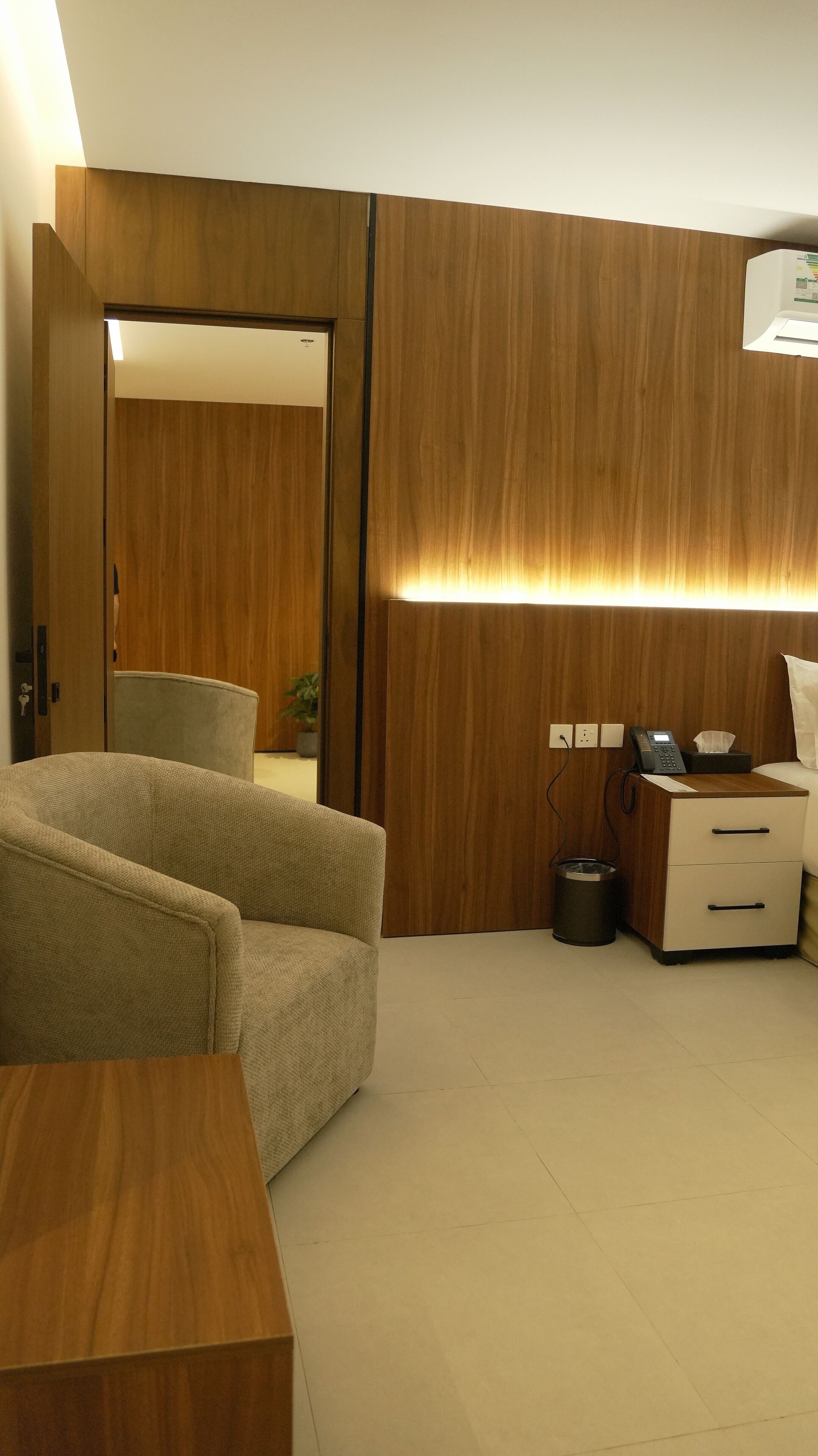 A hotel room with a wooden wardrobe, a sofa, a bedside table, a desk with a telephone, and a trash bin.
