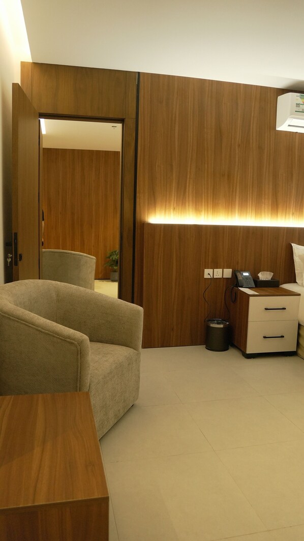 A hotel room with a wooden wardrobe, a sofa, a bedside table, a desk with a telephone, and a trash bin. - Roses S Hotel (Riyadh)