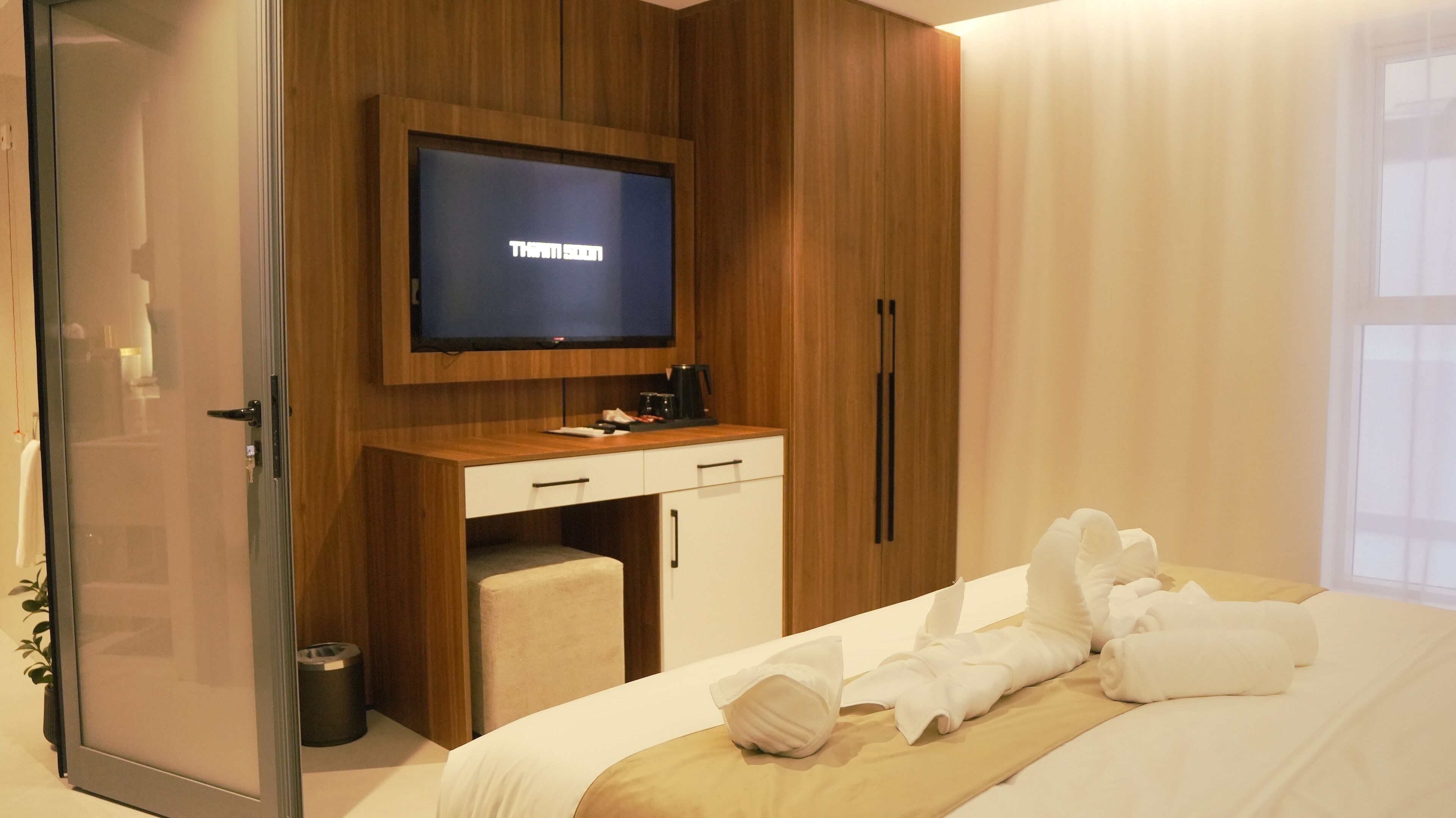 A hotel room with a bed, a television mounted on a wooden wall, a desk with a chair, and a small trash bin.