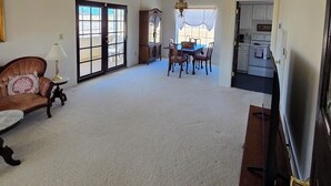 Ein geräumiges Wohnzimmer mit Deckenventilator, ein Essbereich mit Tisch und Stühlen sowie eine durchgehende Tür erkennbare Küche.