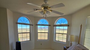 Ein Schlafzimmer mit Bett, Deckenventilator, drei Rundbogenfenstern und Blick auf eine Wüstenlandschaft.