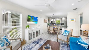 A spacious living area with a dining table, a sofa, and a coffee table. There is a TV mounted on the wall, a ceiling fan, and a kitchen in the background. - Seashells Unit 33 – Relaxing Sanibel Island Beach Retreat (Sanibel)