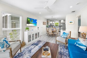 A spacious living area with a dining table, a sofa, and a coffee table. There is a TV mounted on the wall, a ceiling fan, and a kitchen in the background. - Seashells Unit 33 – Relaxing Sanibel Island Beach Retreat (Sanibel)