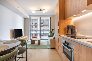 Eine moderne Küche mit Holzausstattung, integriertem Backofen und Kaffeemaschine. Es gibt einen Essbereich mit rundem Tisch und Stühlen, sowie eine Couch im Hintergrund.