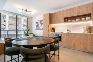 A modern kitchen with wooden cabinets, a round dining table with chairs, and a view of a construction site through large windows. - NEW Penthouse with pool, hot tub, co-working, gym, lounge, games room, BBQ (Miami)