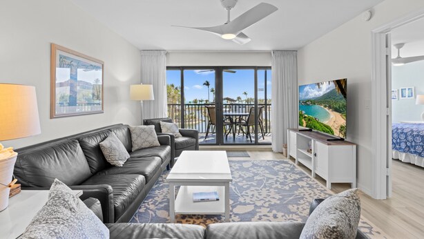 Een moderne woonkamer met een zwarte leren bank, een witte salontafel en een flatscreen-televisie. Er is een balkon met uitzicht op palmbomen en een strand.