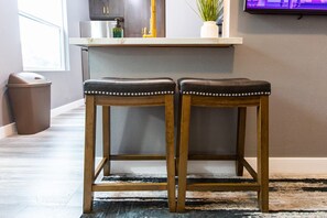 Zwei hölzerne Barhocker mit Metallnieten, ein Mülleimer, ein Blumentopf und eine Küchenarbeitsplatte im Hintergrund.