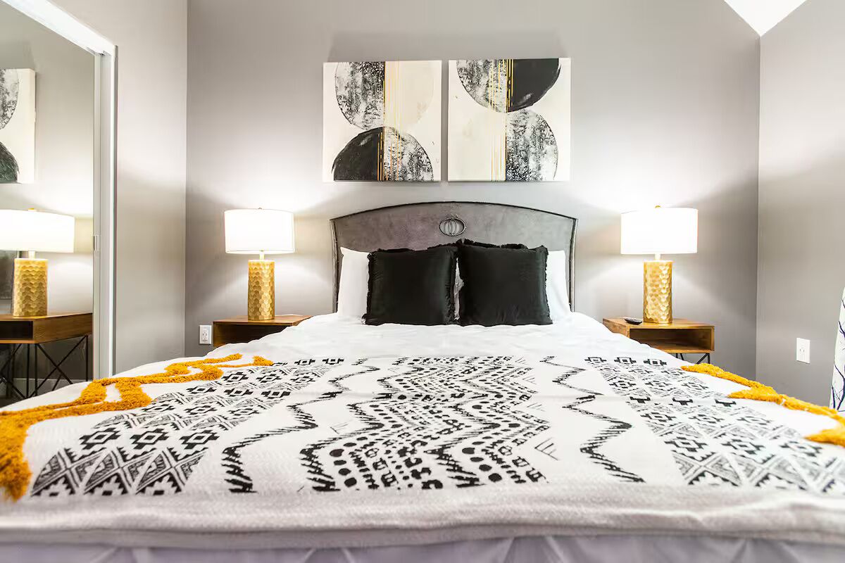 A neatly made bed with a patterned bedspread, two bedside lamps, and abstract wall art above the bed.