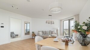 A modern living room with a wooden dining table, white chairs, a sofa, and a large window. - Apartment With Direct sea View (Rewal)