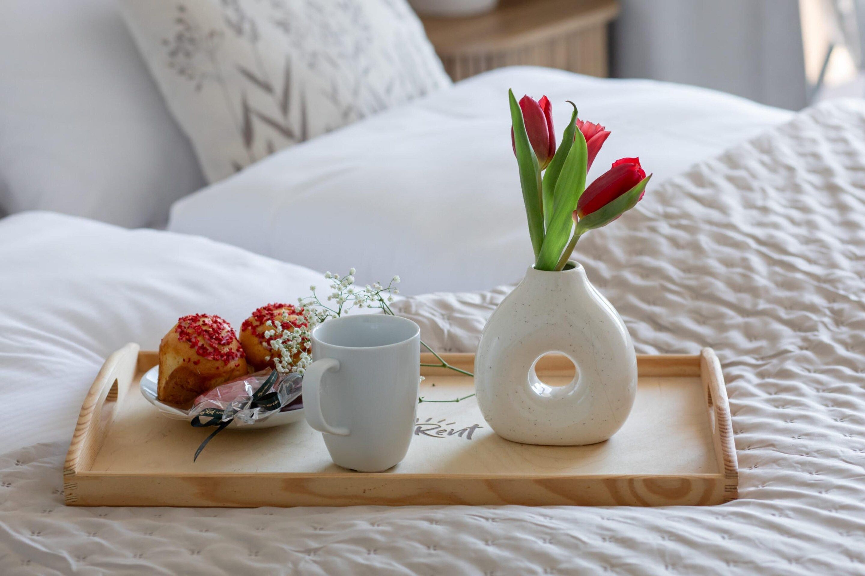 A tray with a mug, a vase with red tulips, and a pastry on a bed with white bedding.