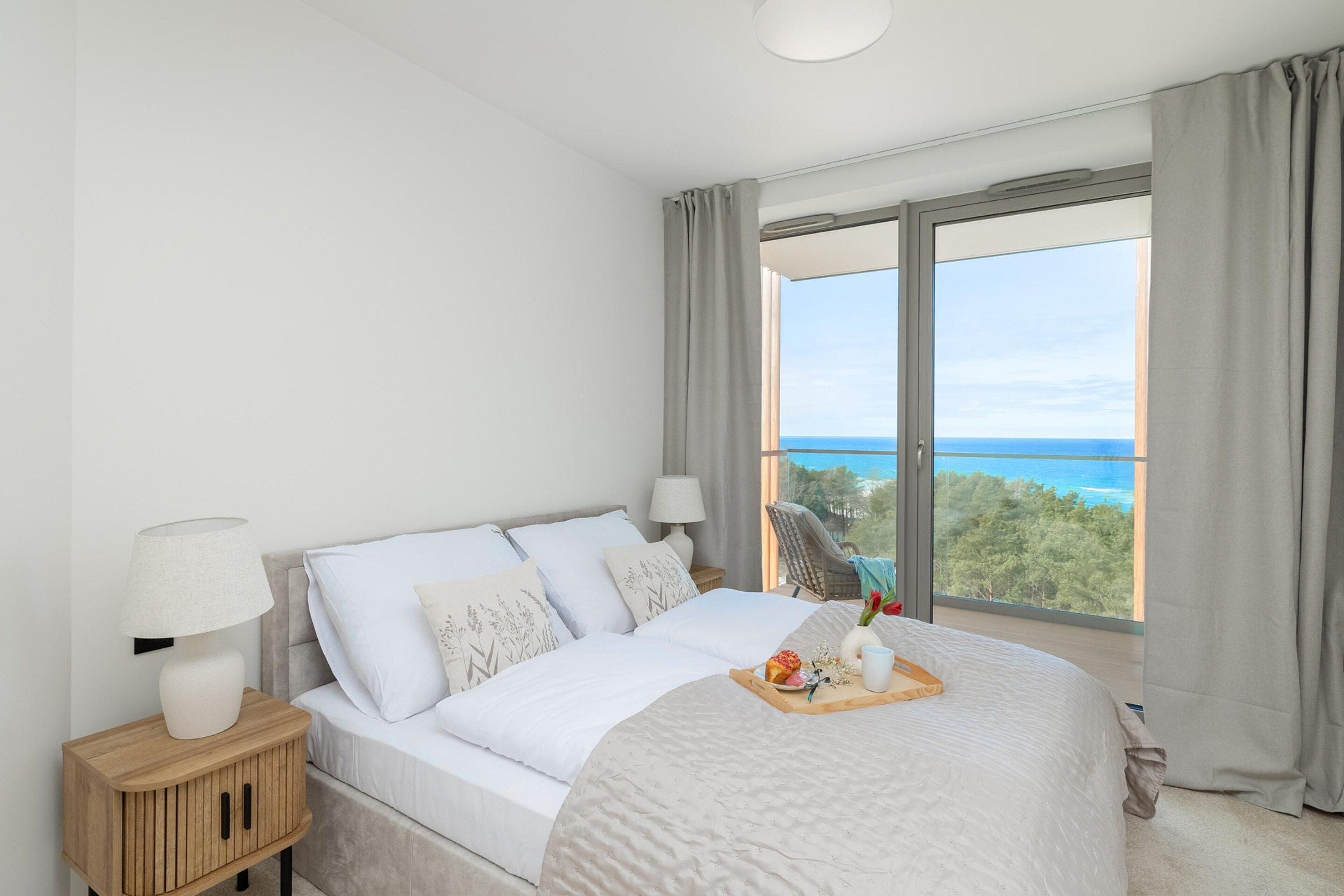 A bedroom with a bed, bedside lamps, a nightstand, and a view of the sea.