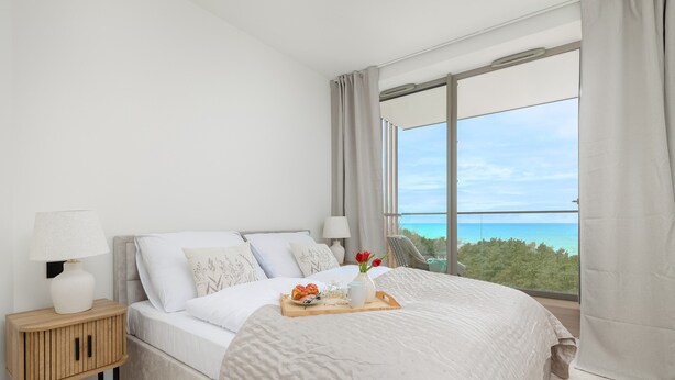 Un dormitorio con una cama grande, una mesita de noche, una bandeja con flores y vistas al mar.