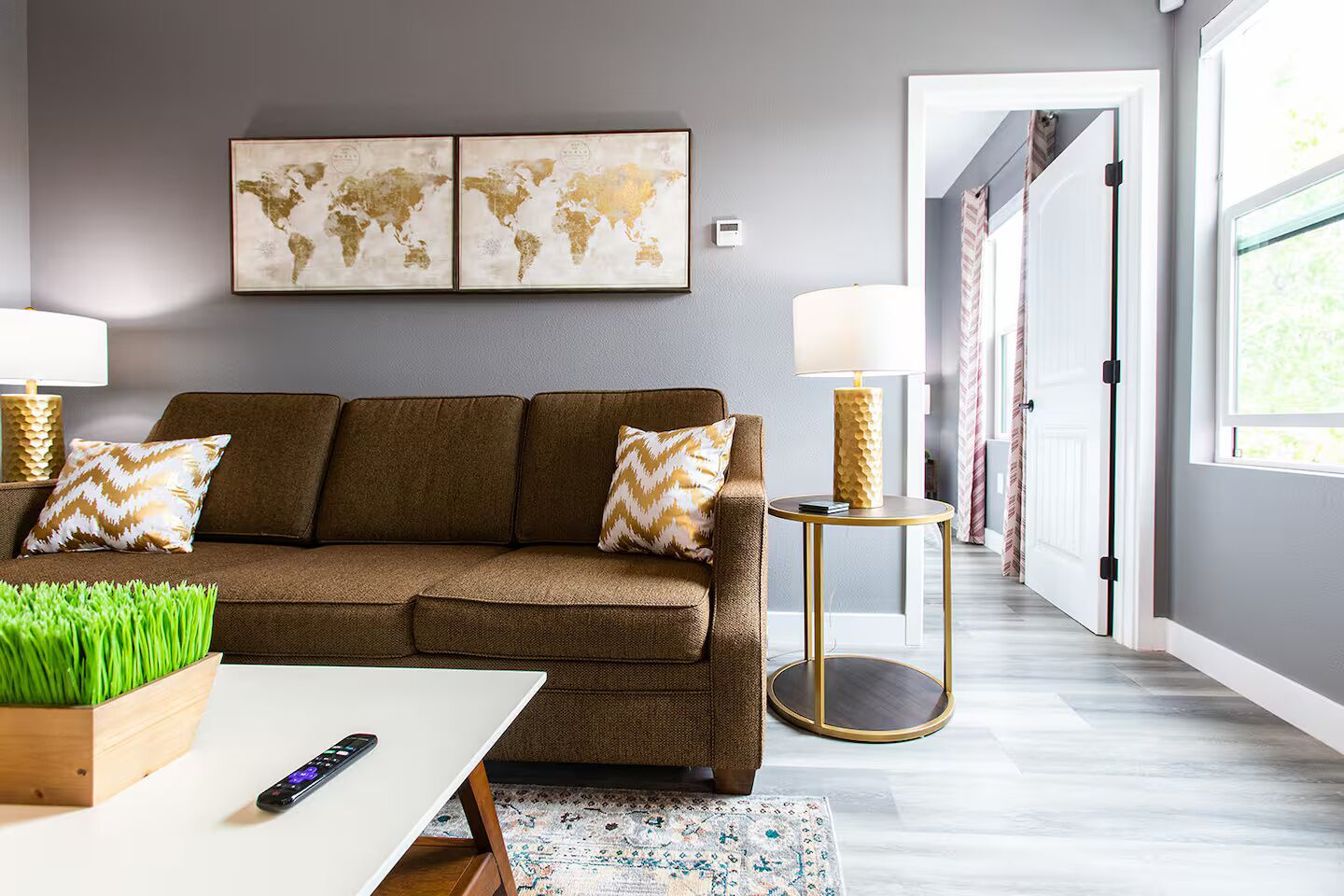 A modern living room with a brown sofa, two decorative pillows, a small round table with a lamp, a coffee table with a remote control, and a world map artwork on the wall.