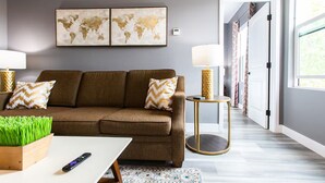Ein modernes Wohnzimmer mit einem braunen Sofa, zwei Zierkissen, einem kleinen runden Tisch mit Lampe, einem Couchtisch mit Fernbedienung und einer Weltkarte als Wandkunst.