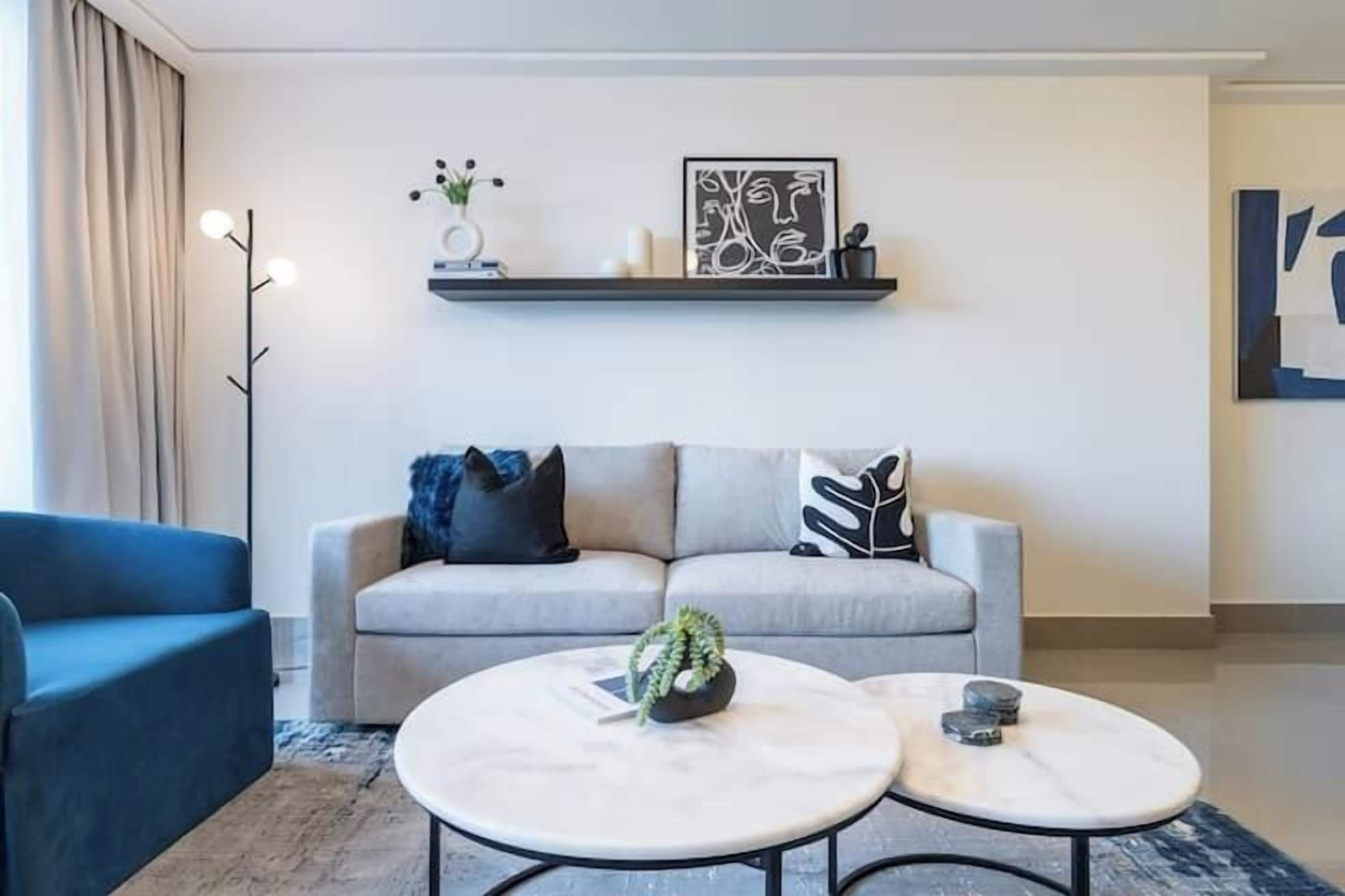A modern living room with a grey sofa, two round tables, a blue armchair, and a wall-mounted shelf with decorative items.