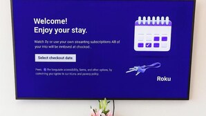 A television displaying a welcome message for Roku, with a calendar icon and a keychain. Below the TV, there is a wooden cabinet with books and a vase with flowers. - Spacious 5BR Retreat Sleeps 13 Premium Bedding Near NRG & Events (Houston)