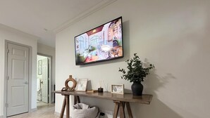 Un couloir avec une console en bois, une plante en pot, un panneau d’accueil encadré et un téléviseur diffusant une image de l’intérieur d’un café.