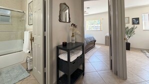 Un couloir mène à une chambre à coucher qui contient un lit, une table de chevet avec un vase, un miroir et une plante en pot.