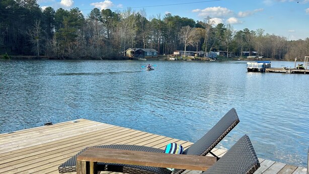 A wooden deck with two lounge chairs and a table, overlooking a calm lake with a dock and houses in the background.