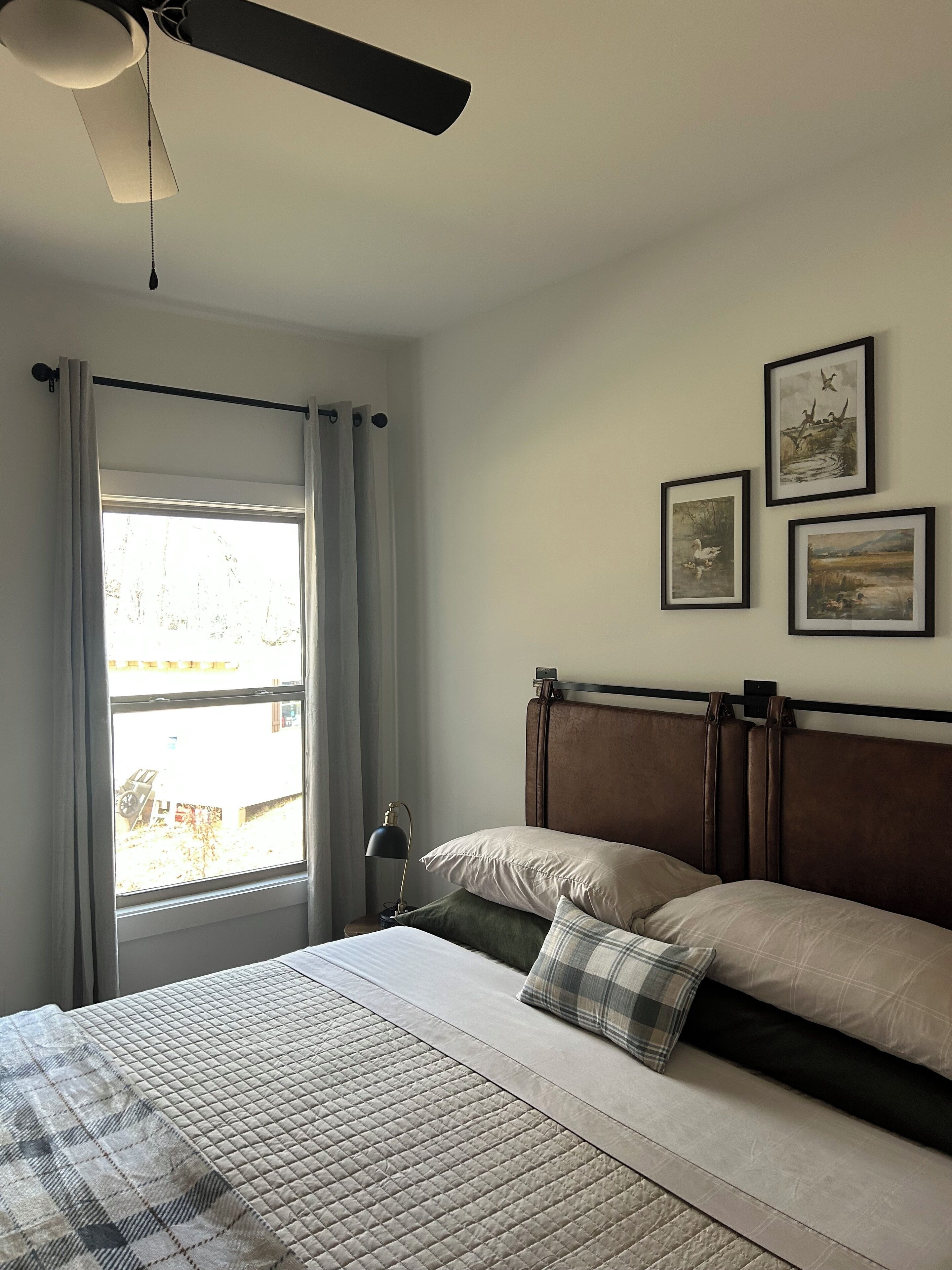 Ein Schlafzimmer mit Bett, Deckenventilator, Fenster mit Vorhängen und drei gerahmten Bildern an der Wand.