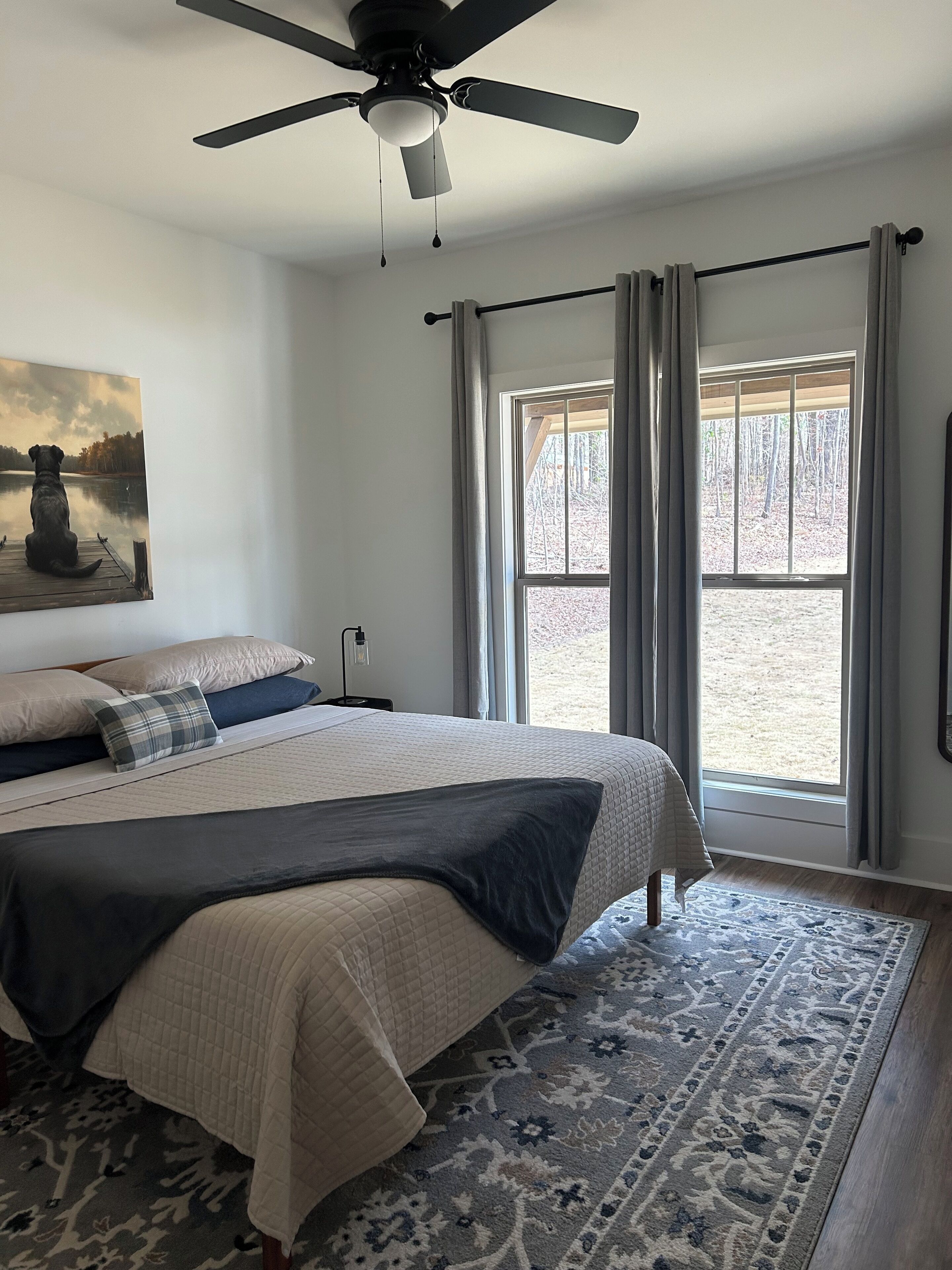 A bedroom with a bed, a ceiling fan, a window with curtains, and a painting on the wall.
