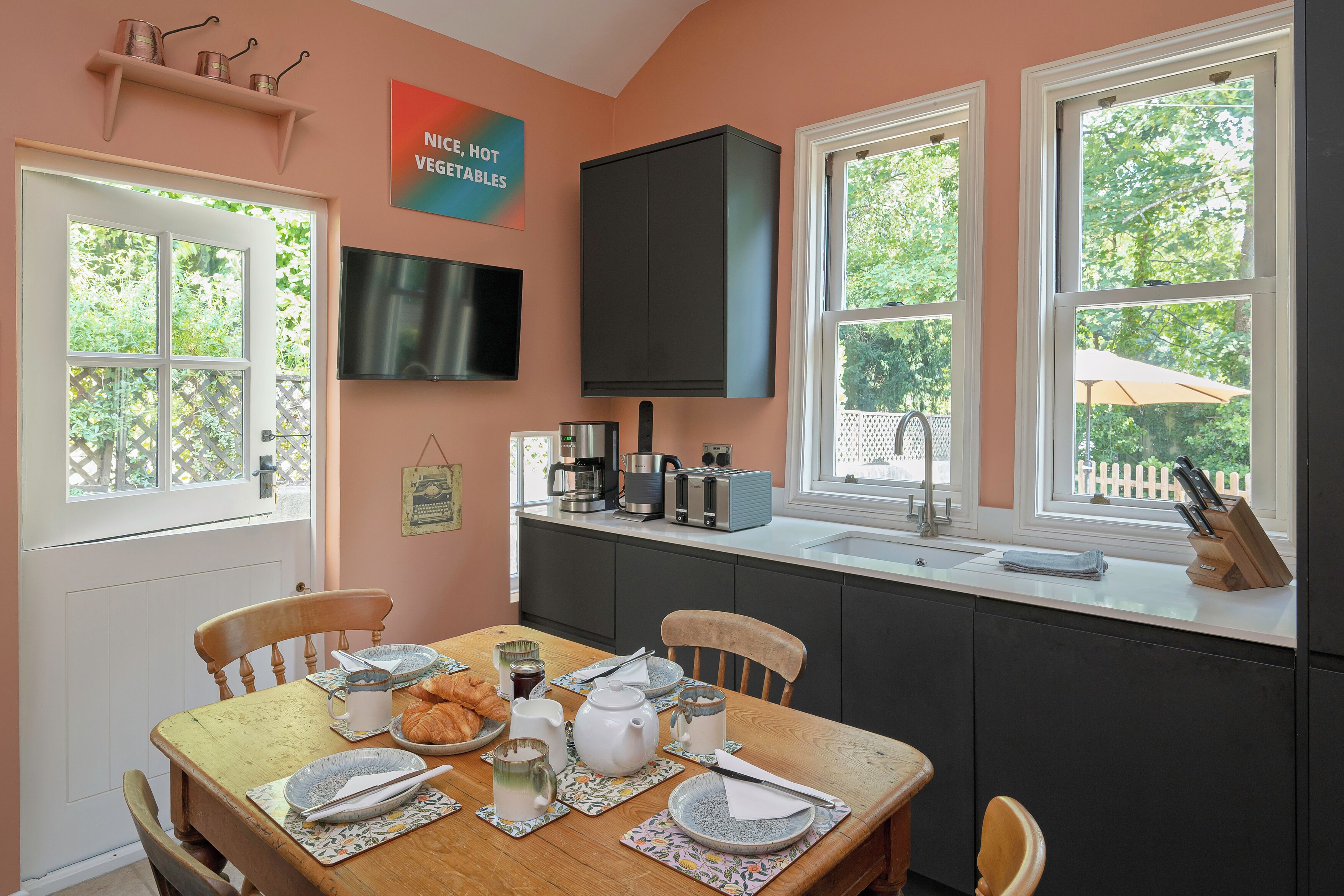 A kitchen with a dining table set for breakfast, a flat-screen TV mounted above the counter, and a window with a view of greenery.