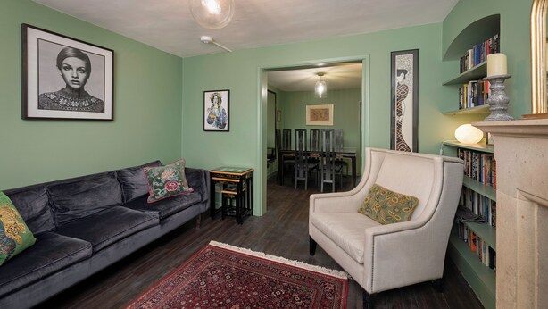 Ein Wohnzimmer mit einem dunkelbraunen Ledersofa, einem hellen Sessel, einem roten Teppich mit Muster und einem Bücherregal mit Büchern.