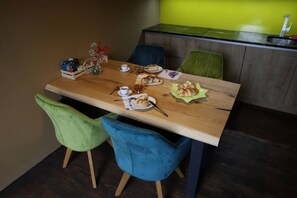 Un comedor con una mesa de madera, dos sillas y una silla verde y azul. Hay un jarrón con flores, un plato con comida y una taza sobre la mesa.