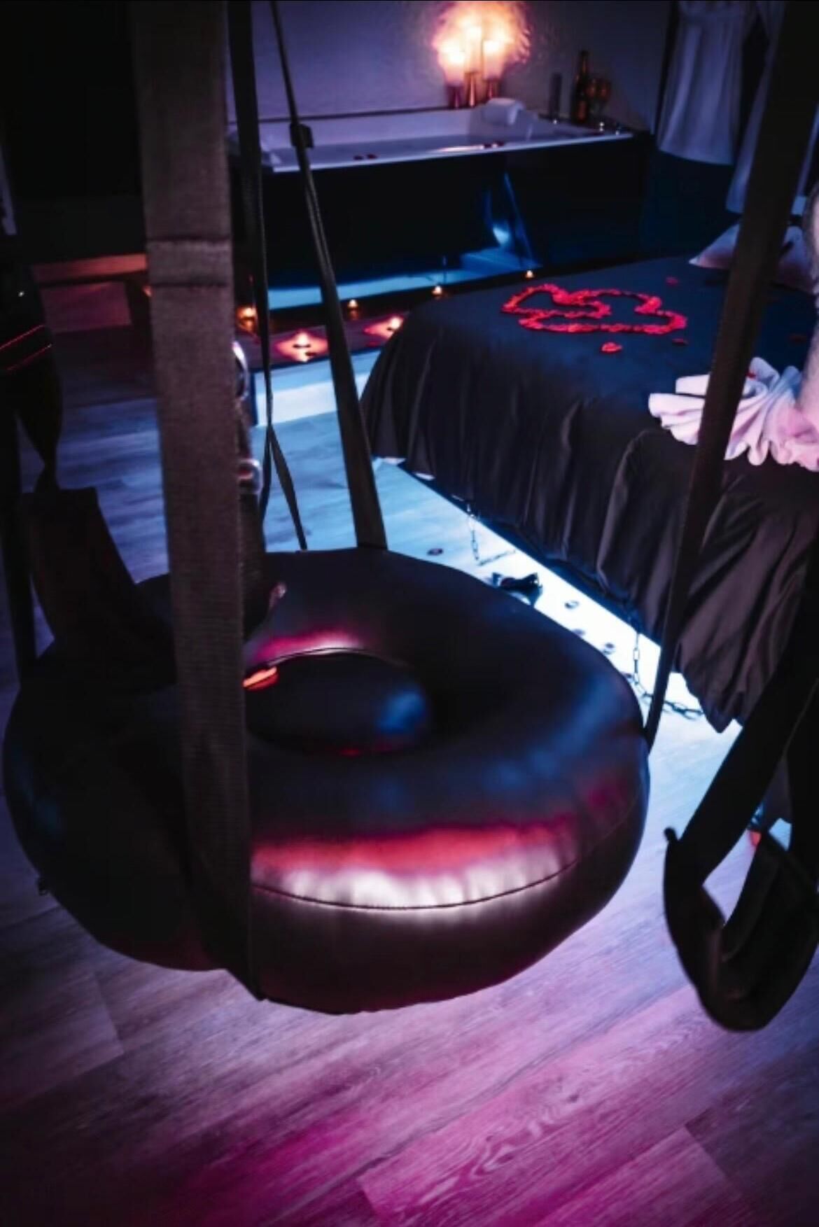 A room with a hanging chair, a bed with a red heart design, and a table with a lamp.