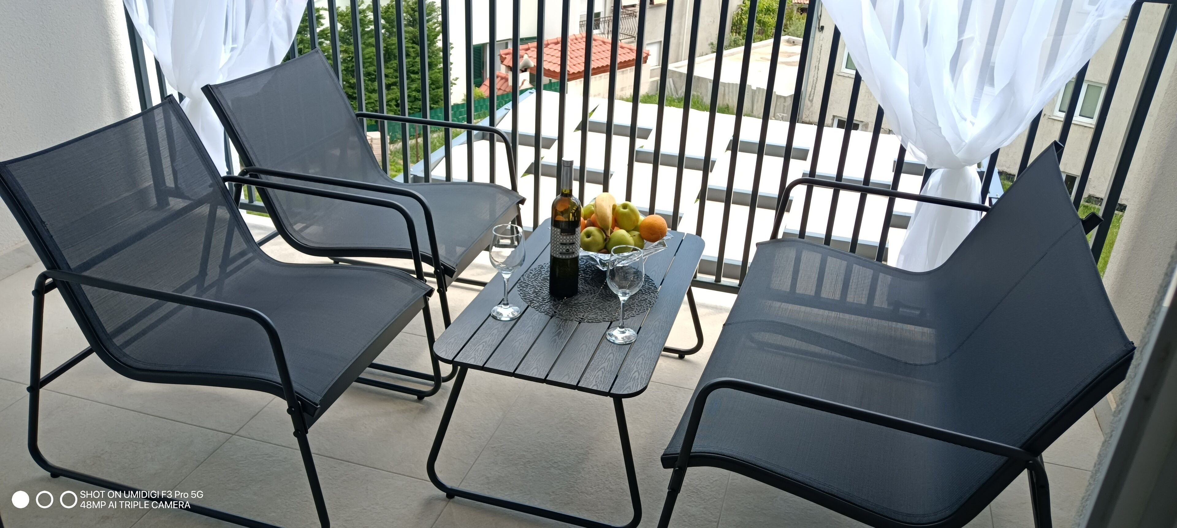 A balcony with two black mesh chairs, a small table with a bottle, a glass, and a bowl of fruit.