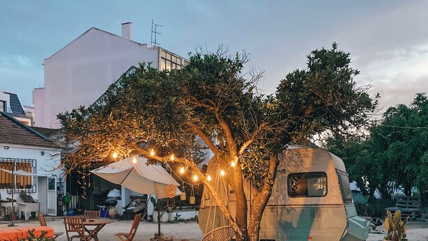 An outdoor seating area with tables and chairs, a tree with string lights, and a camper parked nearby.