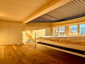 A bedroom with a wooden floor, a bed with a white headboard, a window with a view of the outside, and a skylight with a mesh covering. - Baren Penthouse by Interhome (Zweisimmen)