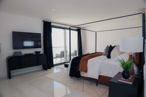 A modern bedroom with a large bed, a TV mounted on the wall, a bedside table with a lamp, and a view of the outdoors through a sliding door.