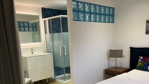 Un dormitorio con cama, mesita de noche, lámpara, baño con espejo y ducha.