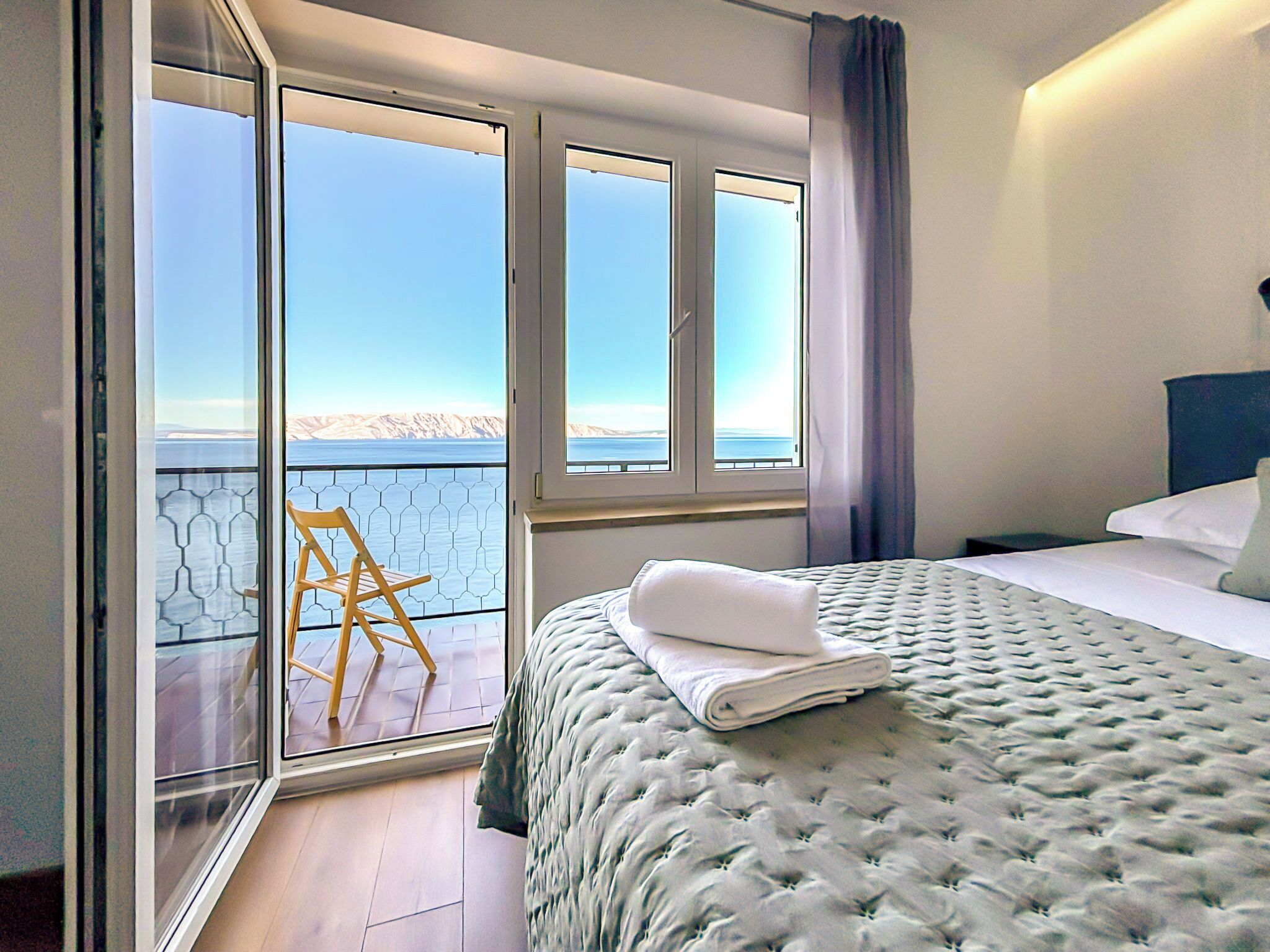 A bedroom with a bed, a chair, and a view of the sea through large windows.